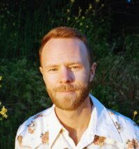 headshot of miles griffis, centered in frame he is wearing a white collared shirt with red bunches of flowers, he is sitting against a green bush outside in a garden area