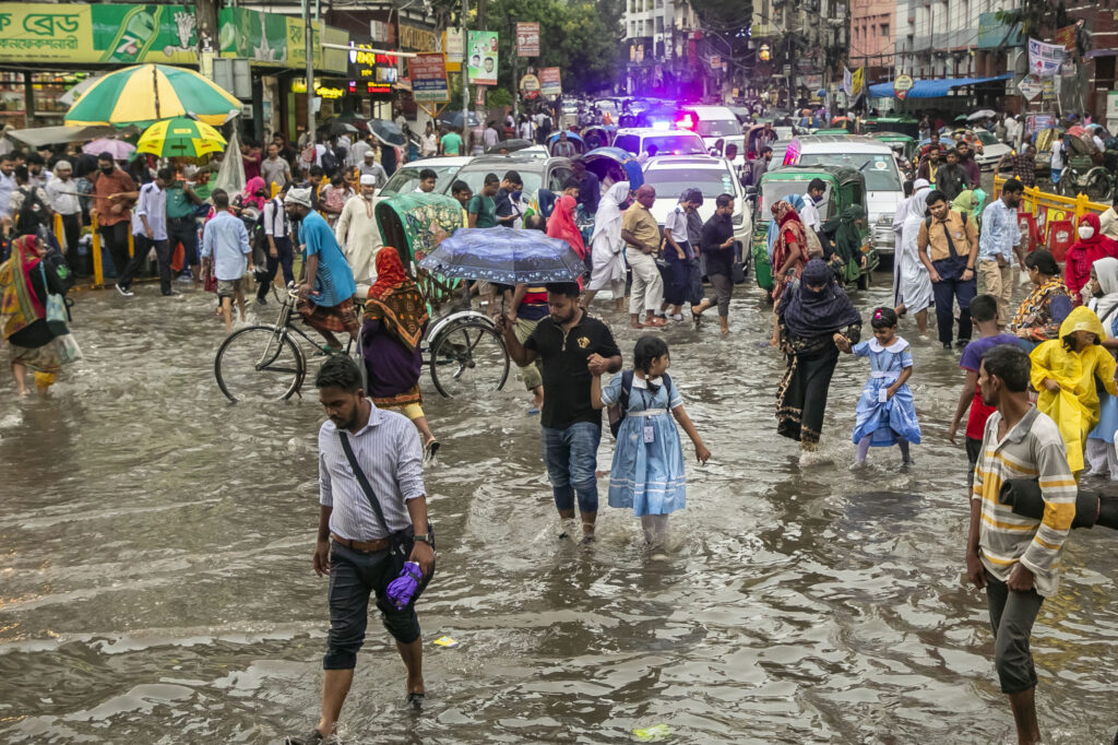 On a cloudy day on a very crowded street, parents holding their children's hands walk through floodwaters almost as high as their knees