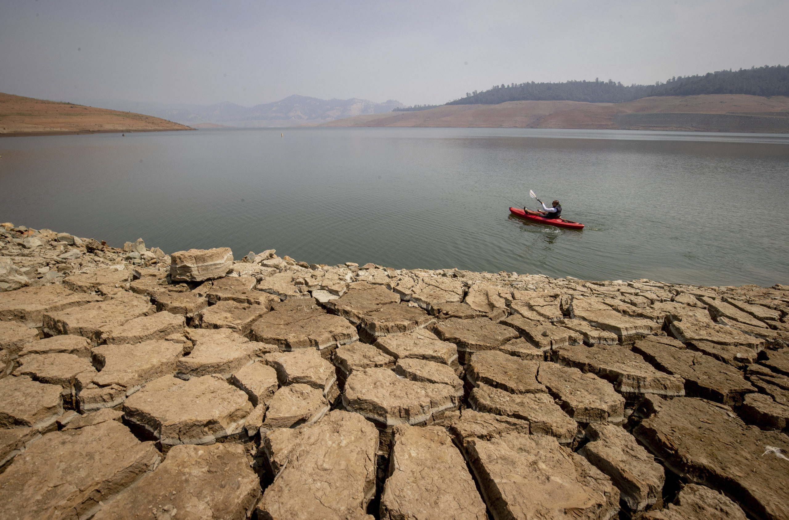 A kayaker paddles on a lake, near the shore, with the mountains in the background on a foggy day. The shoreline, which is rocky, takes up most of the foreground