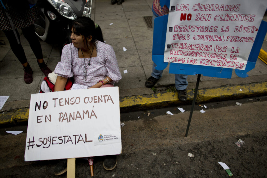 A woman sits on the curb of a sidewalk, holding a sign that reads: "No tengo cuenta en Panama #SoyEstatal." Behind her, another woman holds a sign that reads "-Los cuidadanos no son clientes -Respetaras la diversidad cultural y sexual -Trabajaras por lad incorporación de todos los compañeros"