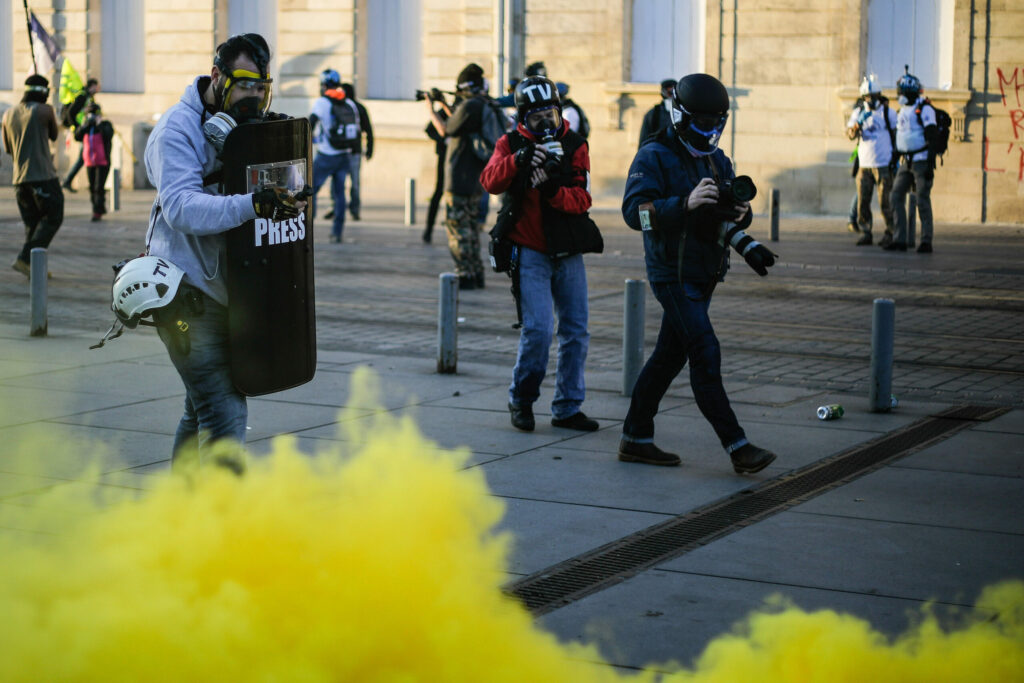 People and journalists walk through a protest in France wearing gas masks and walking past tear gas.