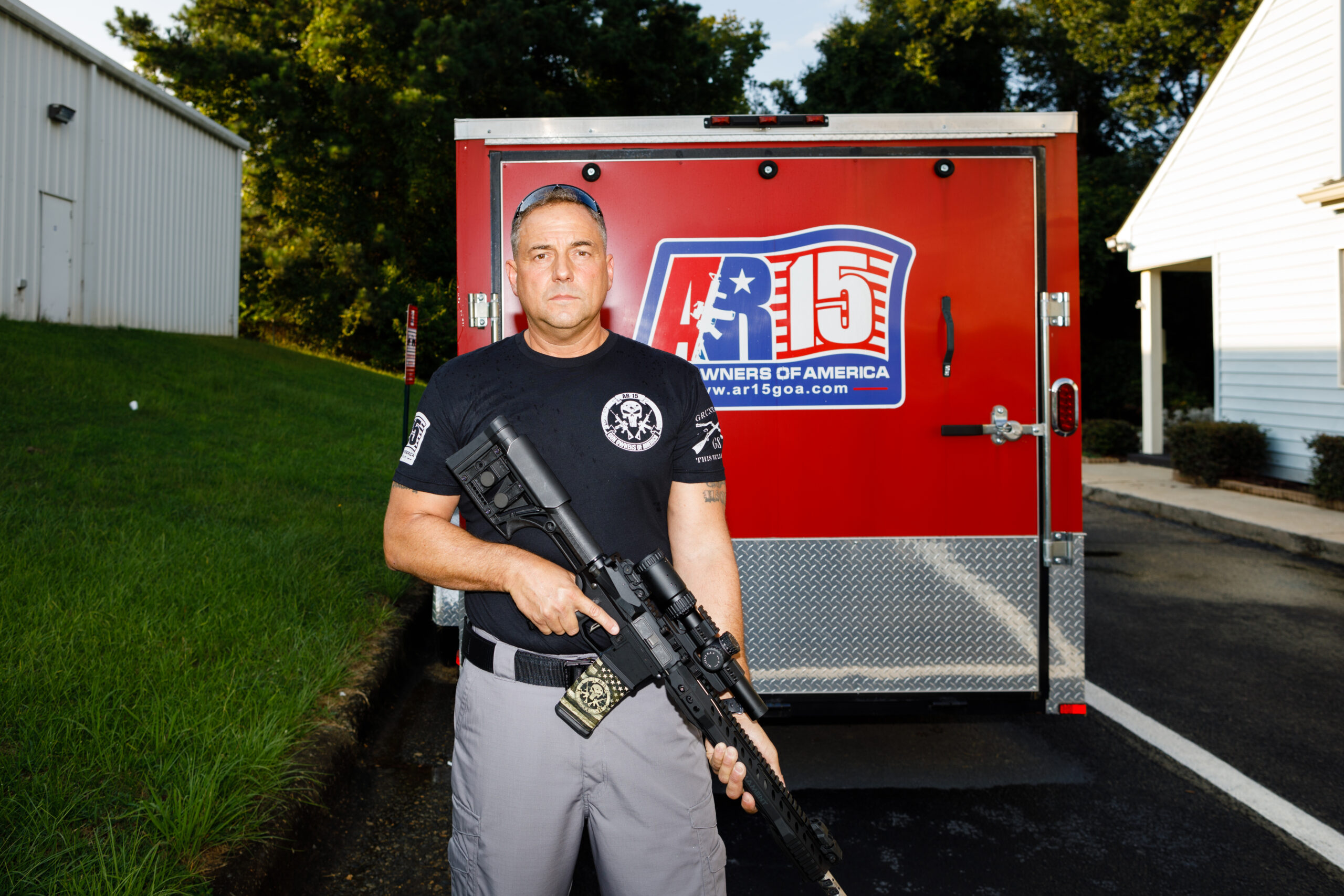 a man is standing outside of his trailer, holding an AR-15 the back of his trailer is red and he is wearing a black t-shirt and gray pants