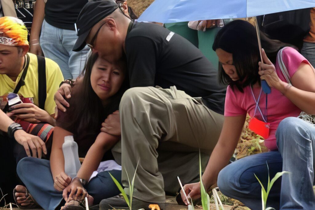 A crowd of people are crouched down on the grass in an outdoor setting. A woman in the center of the picture is leaning into another man's embrace. She is crying and being comforted at the scene.