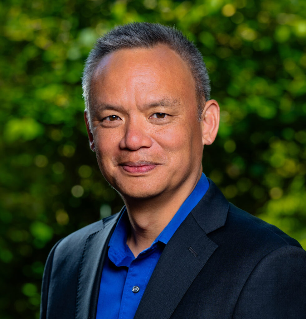 headshot of Edward Wong from the shoulders up, he is against a green leafy background and wearing a bright blue collared shirt and a black blazer