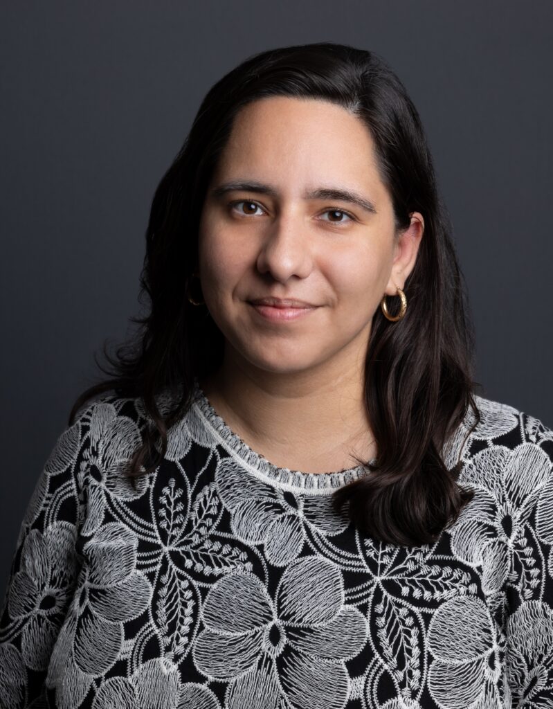 Professional headshot of journalist Nicole Foy from the shoulders up. She is wearing a gray blouse with a black floral pattern against a gray background.