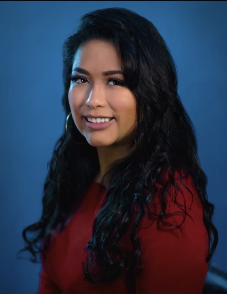 professional headshot of journalist Ximena Bustillo, she is turned to the right and is wearing a dark red blouse against a deep blue background