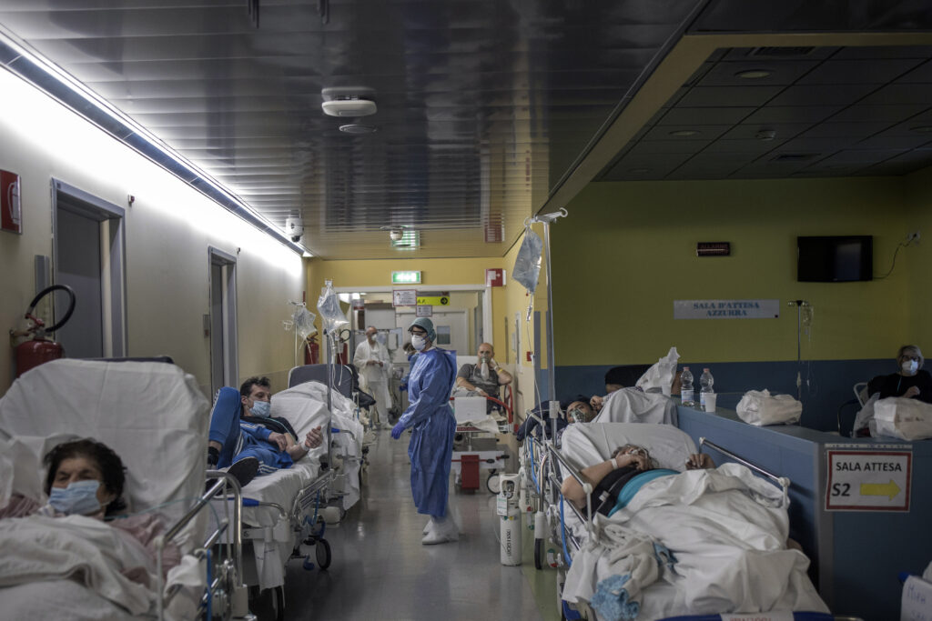 ER department crowded with many patients lying on stretchers, with many doctors and medical staff in the room wearing PPE. 