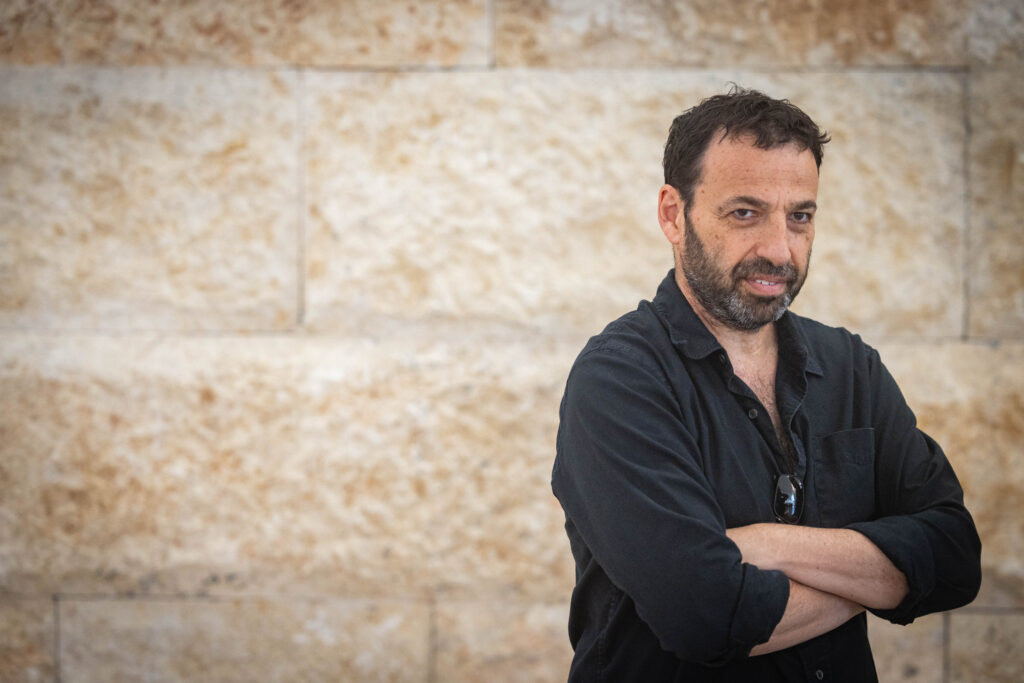 raviv drucker in a black button down posing against a stone sandy brown wall
