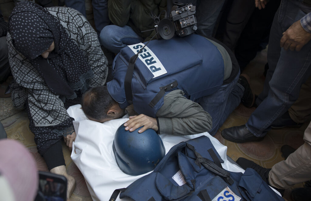 mourners kneel over samer abu daqqa's body, wrapped in a white sheet, at his funeral. a safety helmet is placed over his body.