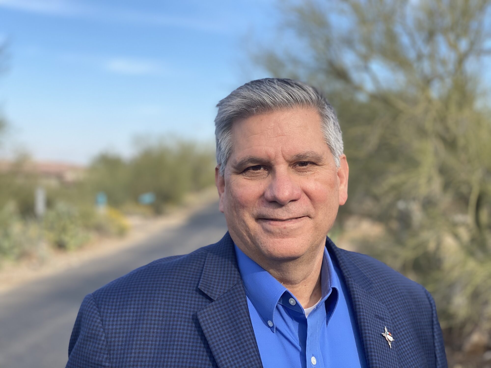 Mark Trahant headshot, shoulders up, wearing a bright blue button up shirt and a navy blue jacket. The desert road and blue sky is behind him.