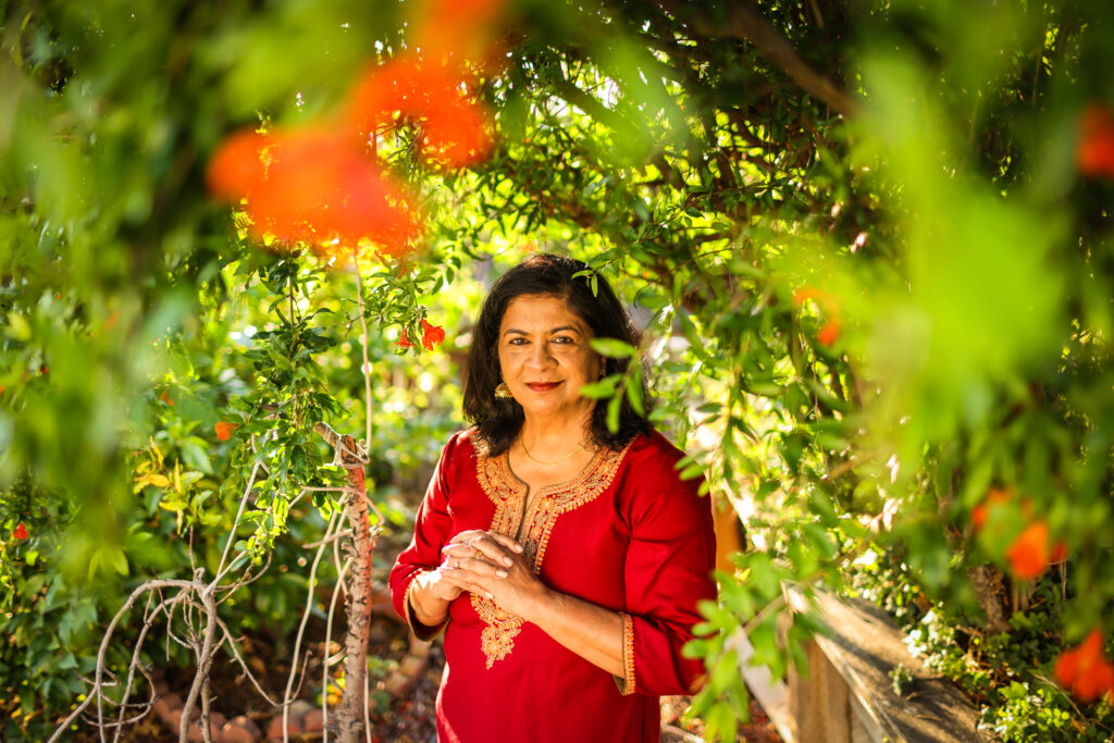 Vandana wears a flowing crimson tunic and stands underneath a canopy of orange flowers with lime green leaves