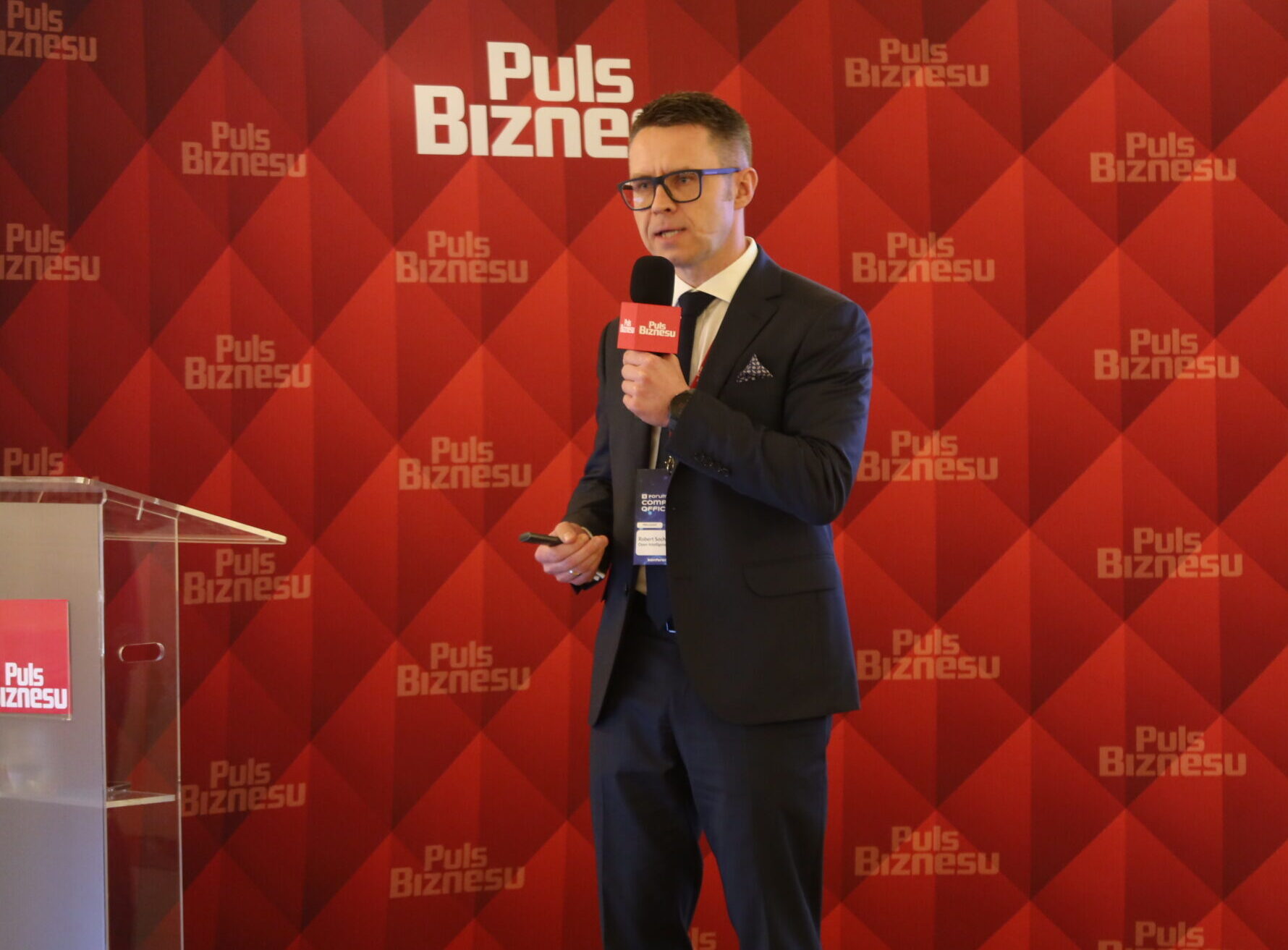 man stands behind a red background at a conference, he is wearing a black suit and white button down
