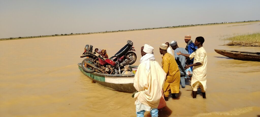 a group of villagers approach a wooden boat carrying people and a motorcycle 