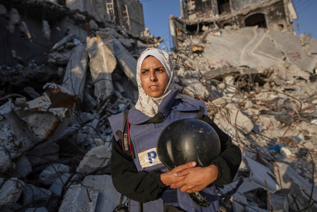 woman stands in front of rubble in gaza, wearing a blue press vest and white hijab