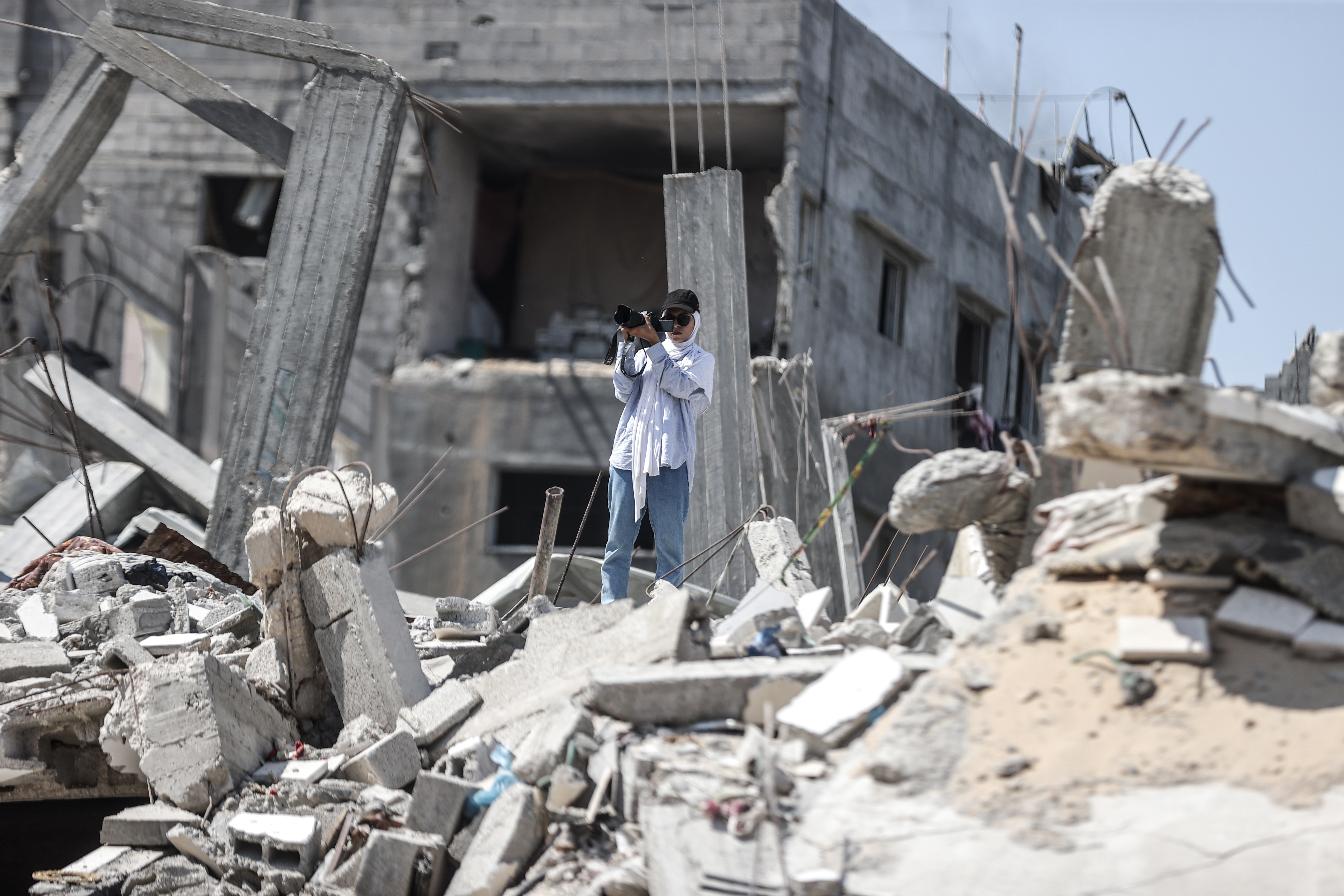 woman stands in blue jeans and a shirt on top of rubble in Gaza