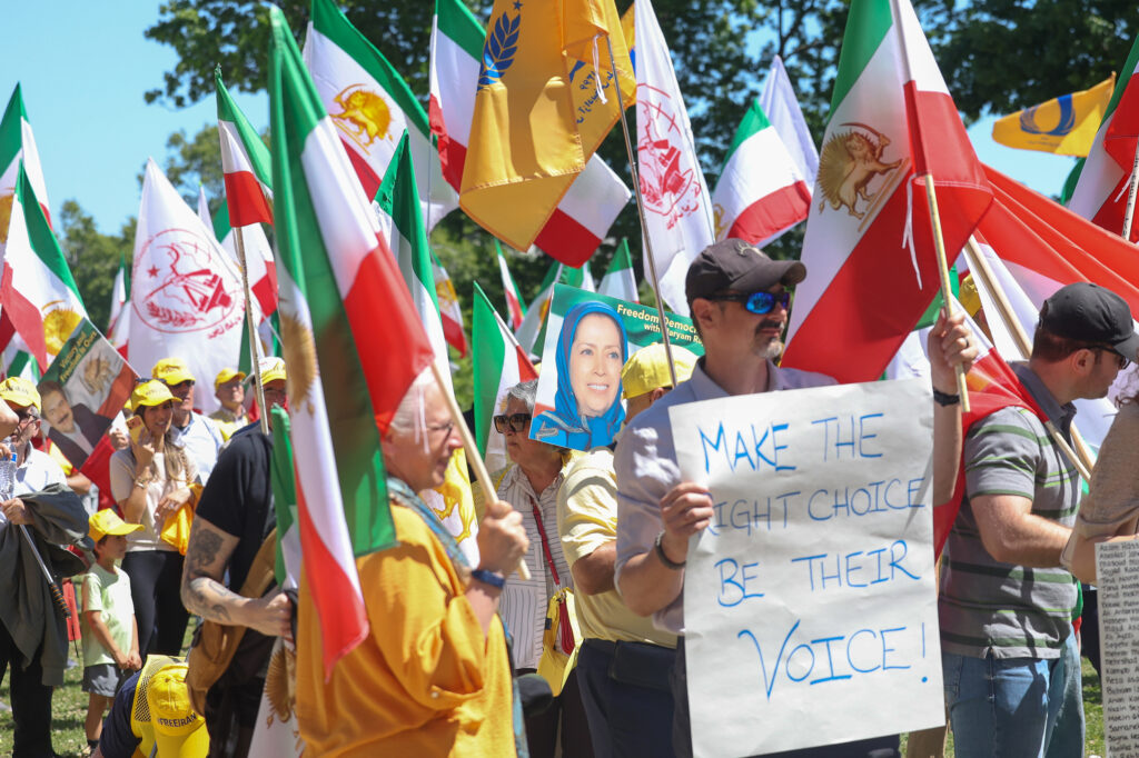rally goers wave iranian flags on a sunny day in dc. a man wearing early 2000s sleek sunglasses and a baseball cap holds up a sign to the camera, "make the right choice, be their voice!"