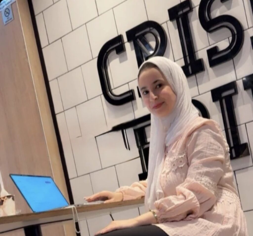 Athar, the article's author, works from a brightly lit cafe. the walls have white tiles and the furniture is a light brown color, very modern. athar is looking at the camera and seated at a table, with one hand on a laptop