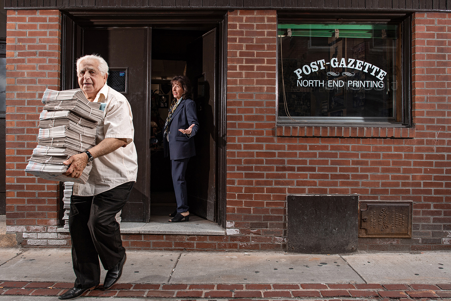 an elderly man with white hair walks outside of a brick building, carrying a stack of freshly printed newspapers. his girlfriend is wearing a pantsuit and is standing behind him, in the doorway of the building.