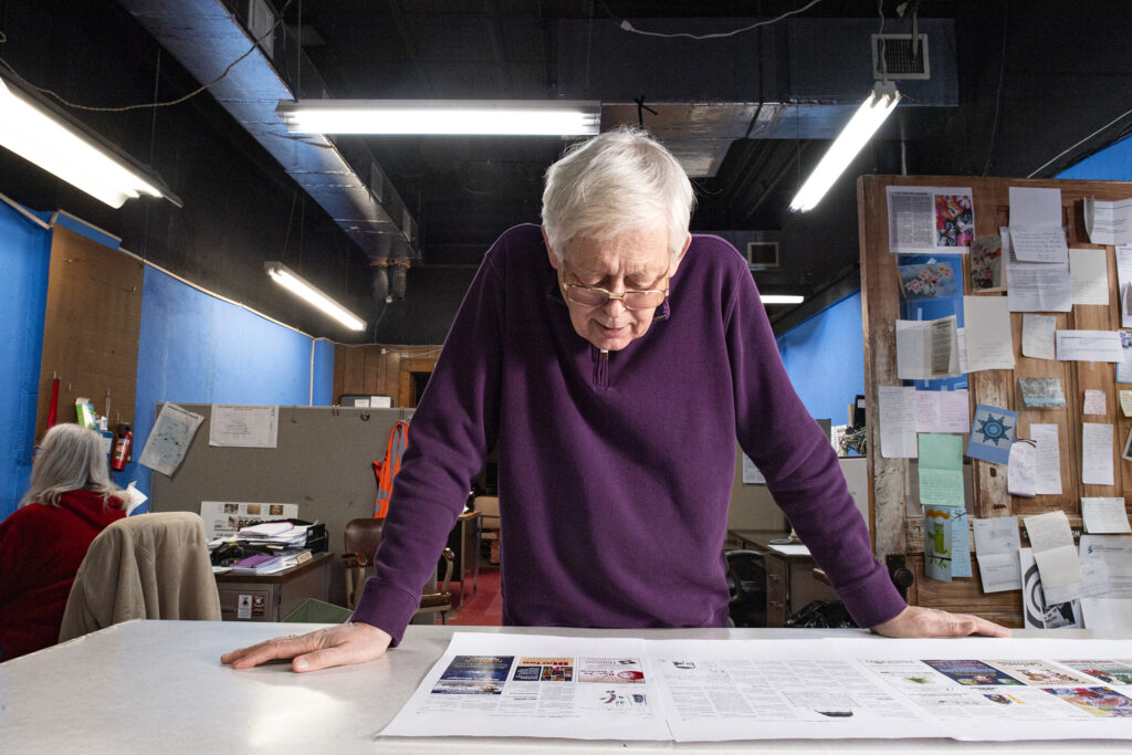 an elderly man wearing a plum sweater leans over a desk, looking down at a newspaper spread
