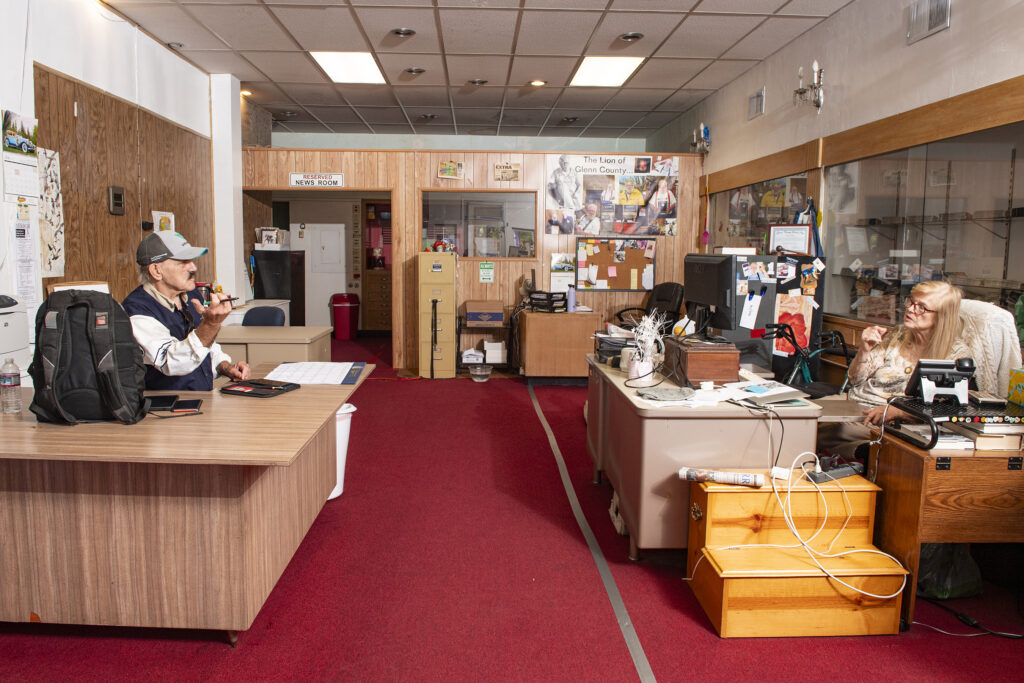 an old newsroom with brown desks and red, vintage carpet. two staff members at opposite sides of the room are talking to each other.