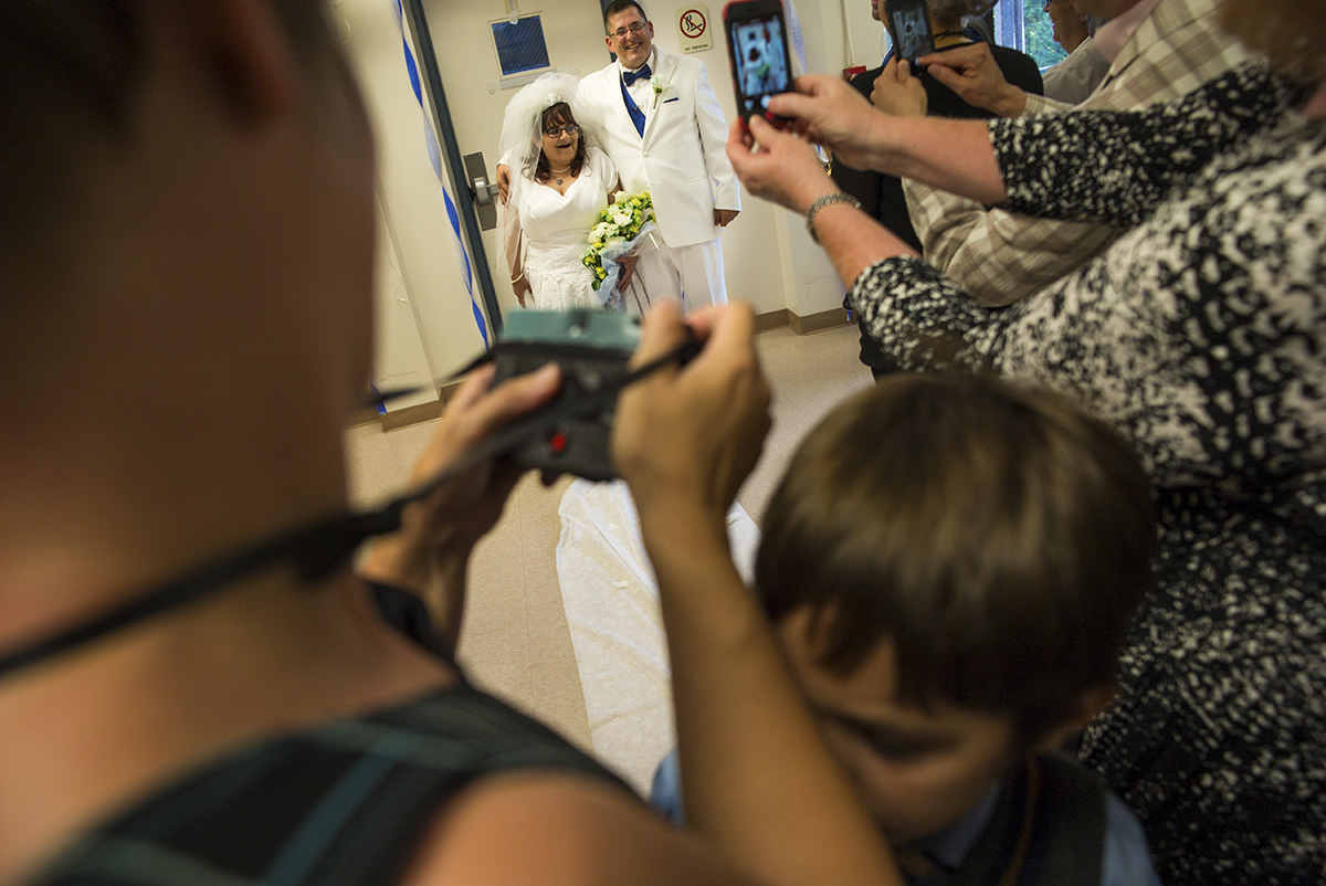 A woman in a white wedding dress holding flowers stands with her husband in a white suit with a blue vest. Friends and family in the foreground take photos of the couple on their wedding day.