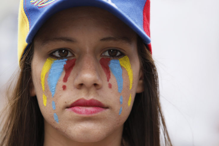 Más de 60 personas han sido asesinadas en la última ola de protestas en Venezuela esta primavera, y cientos más—incluyendo periodistas—han resultado heridos. Una mujer con el rostro pintado con los colores de la bandera nacional de Venezuela asiste a una protesta silenciosa en homenaje a los caídos.