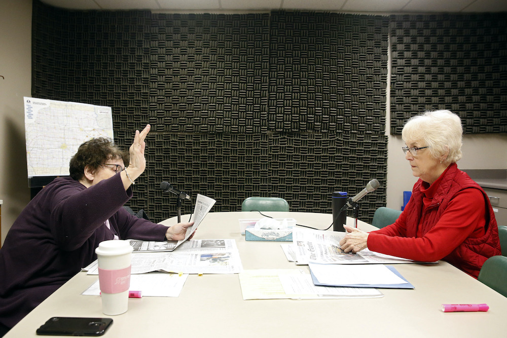 A woman raises her hand to signal she’s nearing the end of an article. A second woman prepares to begin the next as they read a newspaper aloud.