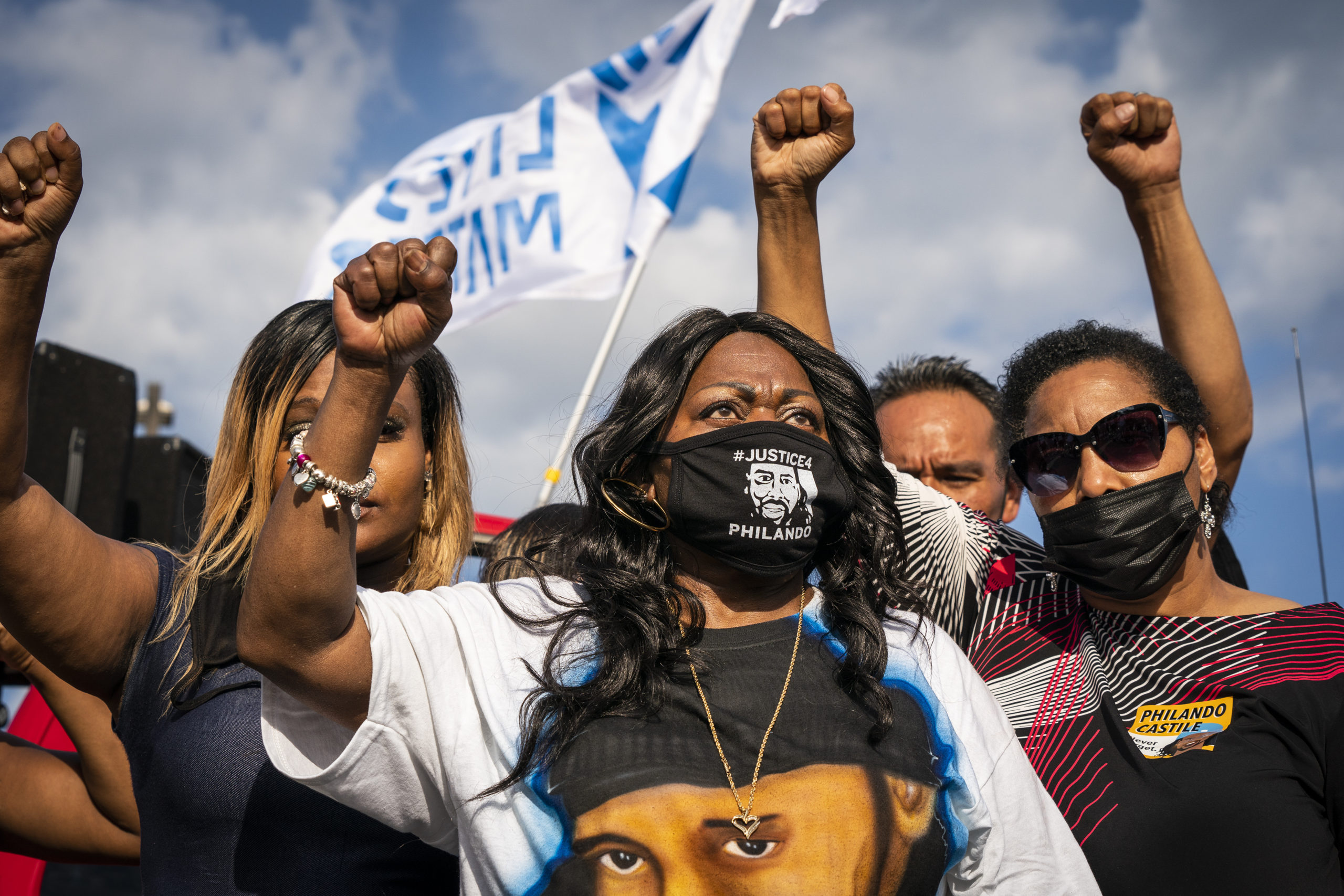 Wearing a mask that says "#Justice4Philando," Valerie Castile and three others raise a fist. A blue and white flag that reads "Black Lives Matter" waves in the background
