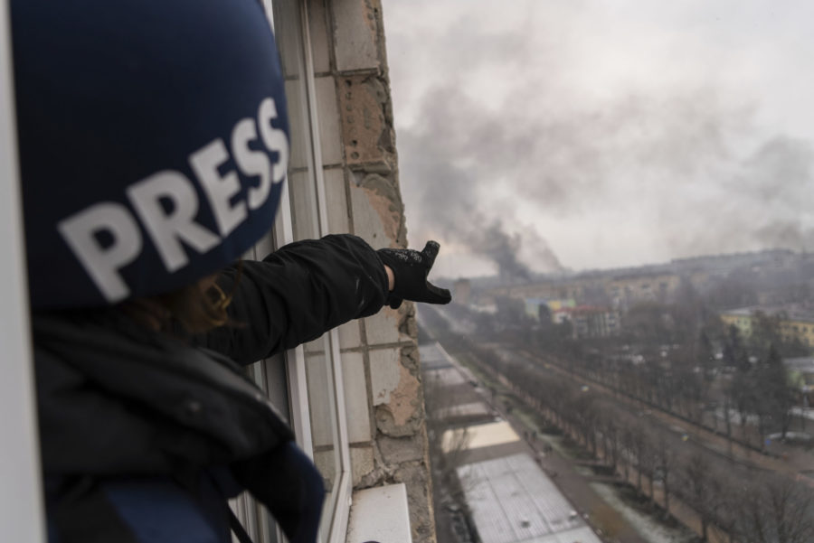 A reporter with a helmet that reads "PRESS" on it hangs out of a window and points a cloud of smoke in the distances