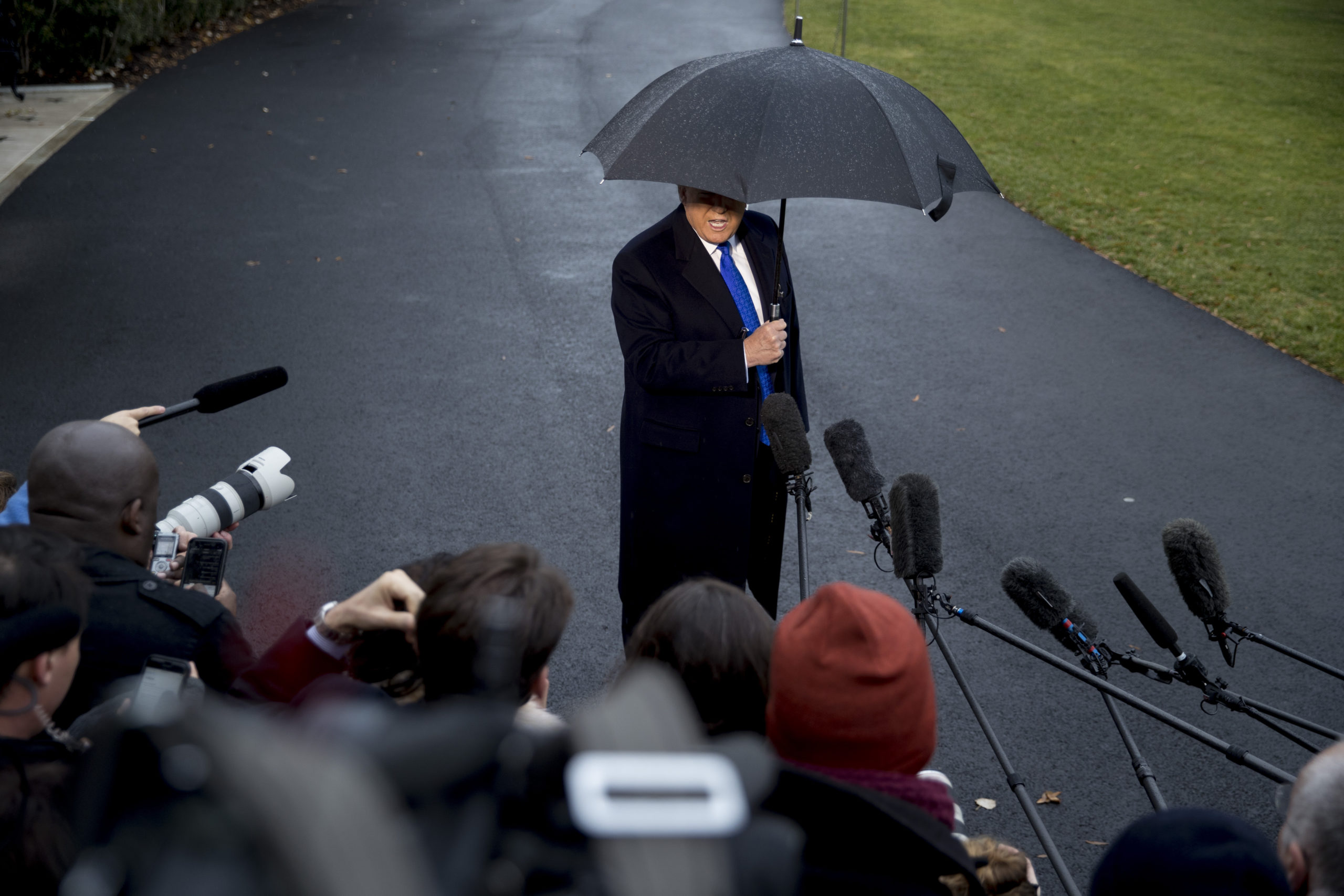 President Donald Trump looks into a crowd of media reporters, with their microphones extended out at him. It is gloomy, and Trump is sporting an umbrella that covers much of his face