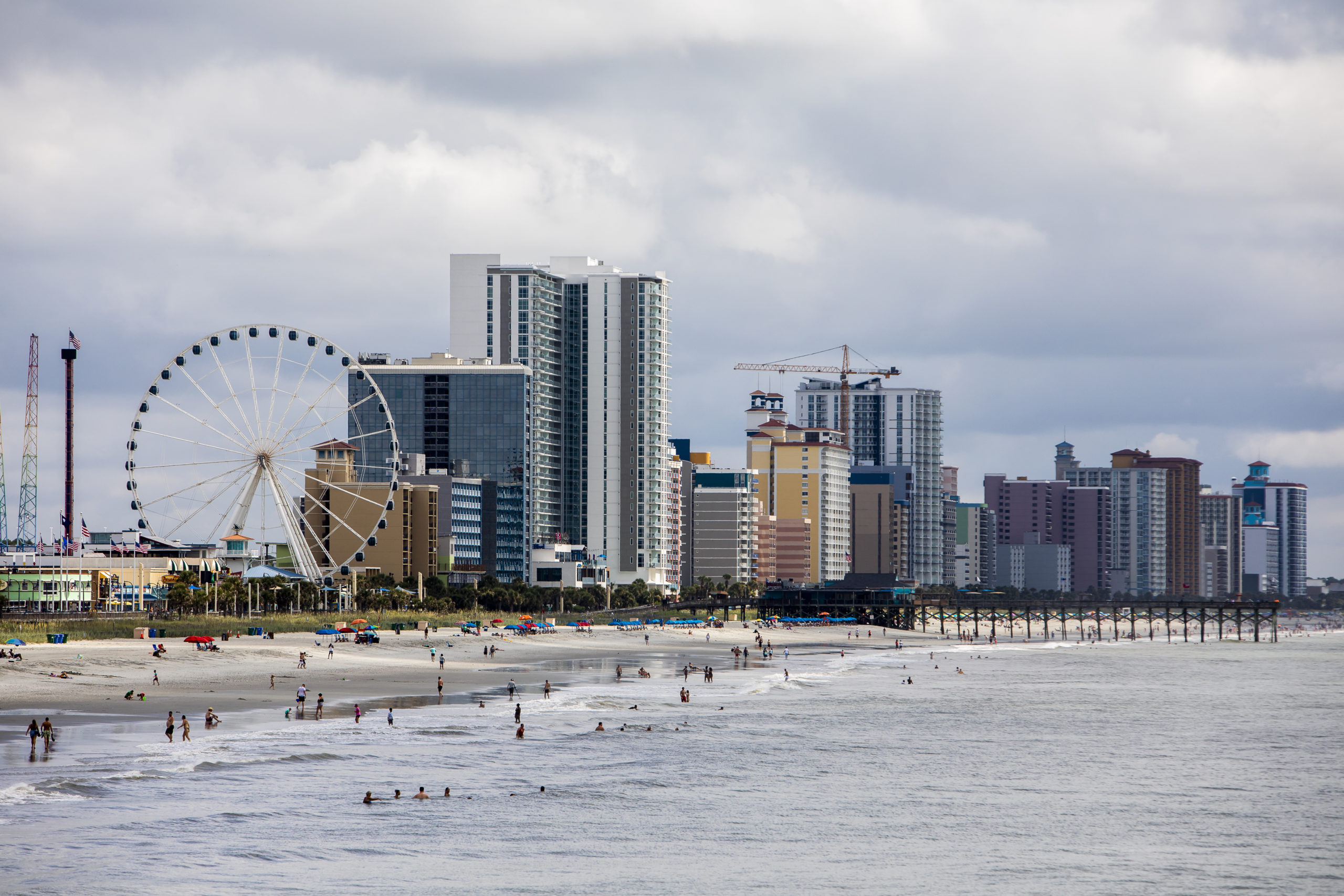 A landscape photo of the beach in Myrtle Beach, South Carolina. To the left of the coastline, there is a ferries wheel in the distance. To the right, tall buildings compose of the skyline. Beachgoers can be seen on the shore.