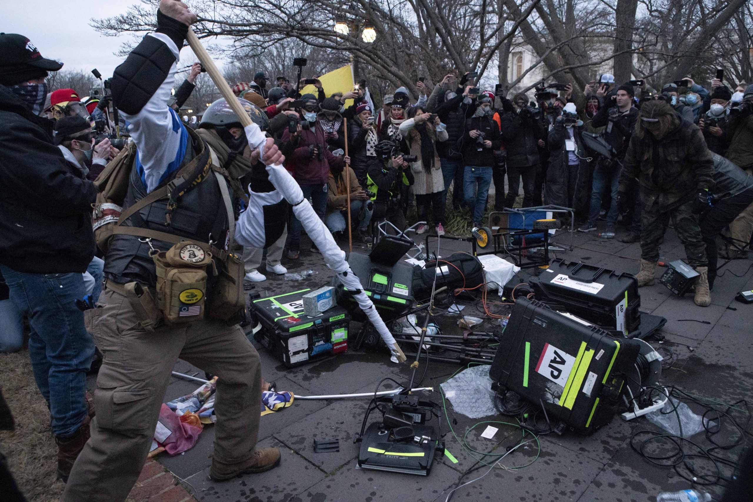 A circle of people gather around a pile of destroyed Associated Press TV equipment. To the left, a man in a helmet, a vest, and camo gear strikes a long wooden pole into the equipment