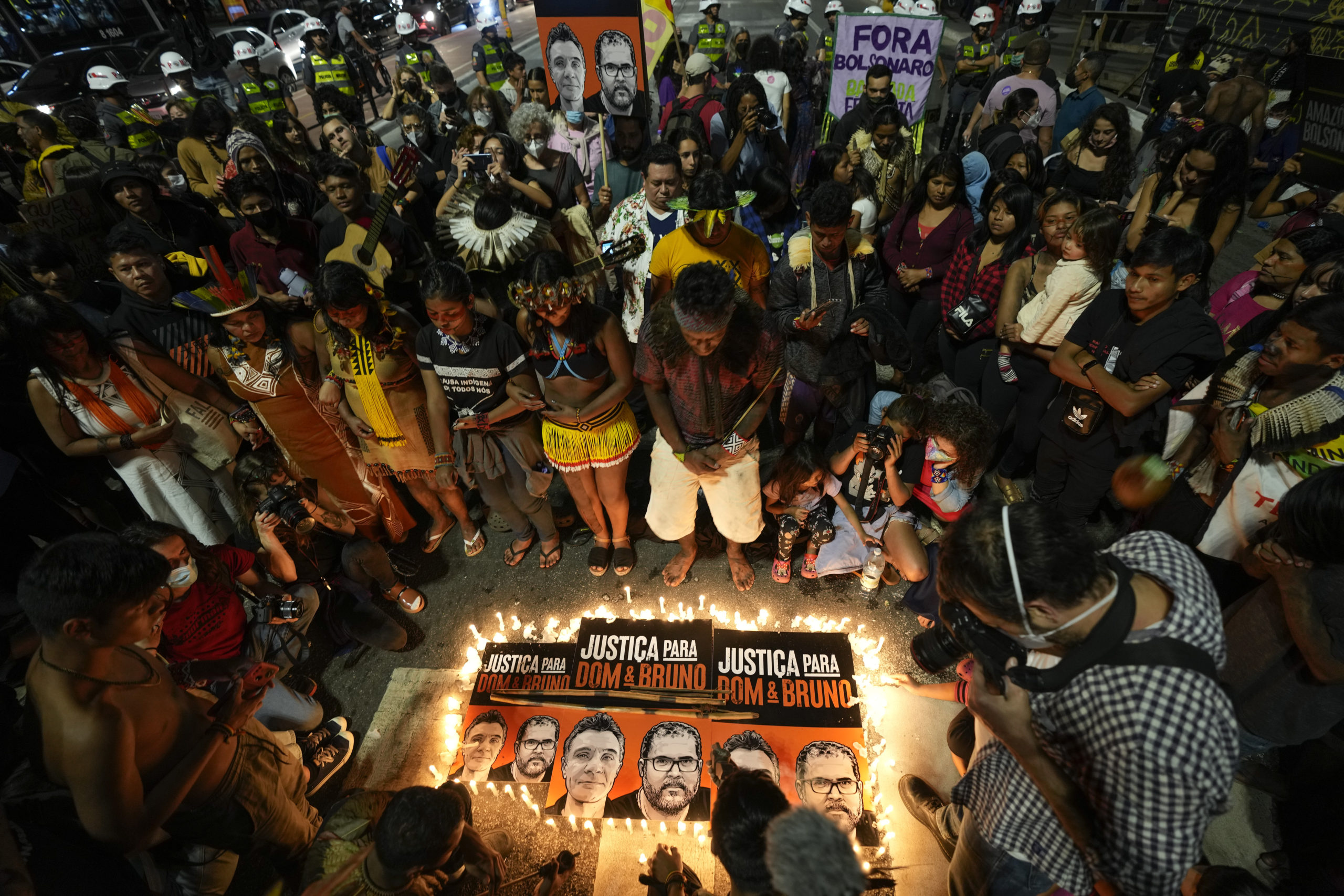 A crowd of Indigenous people gather around a memorial for Dom Phillips and Bruno Pereira. The memorial has three posters, each with Dom and Bruno's face on it, reading "Justiça Para Dom e Bruno" (Translates to Justice for Dom and Bruno), and is surrounded by candles. Toward the back of the crowd, someone holds an enlarged poster of Dom and Bruno; someone else holds a banner that says "Fora Bolsonaro" (Bolsonaro Out).Behind the crowd of Indigenous memorial goers, police wearing helmets and green vests surround them.