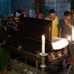 Two men, one in a yellow shirt and one wiping off tears from his eyes, look into the candle-lit casket of Juan Carlos Muniz