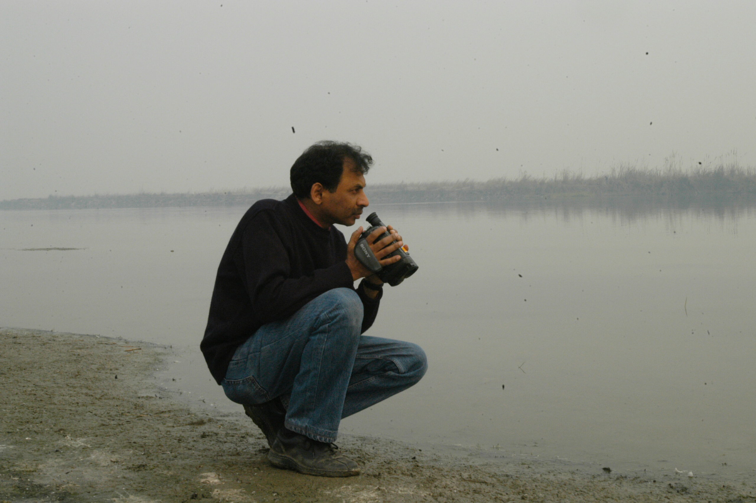 With a grey, cloudy landscape of a lake in the foreground, Ravi Agarawl crouches down over the water with a camcorder in his hands