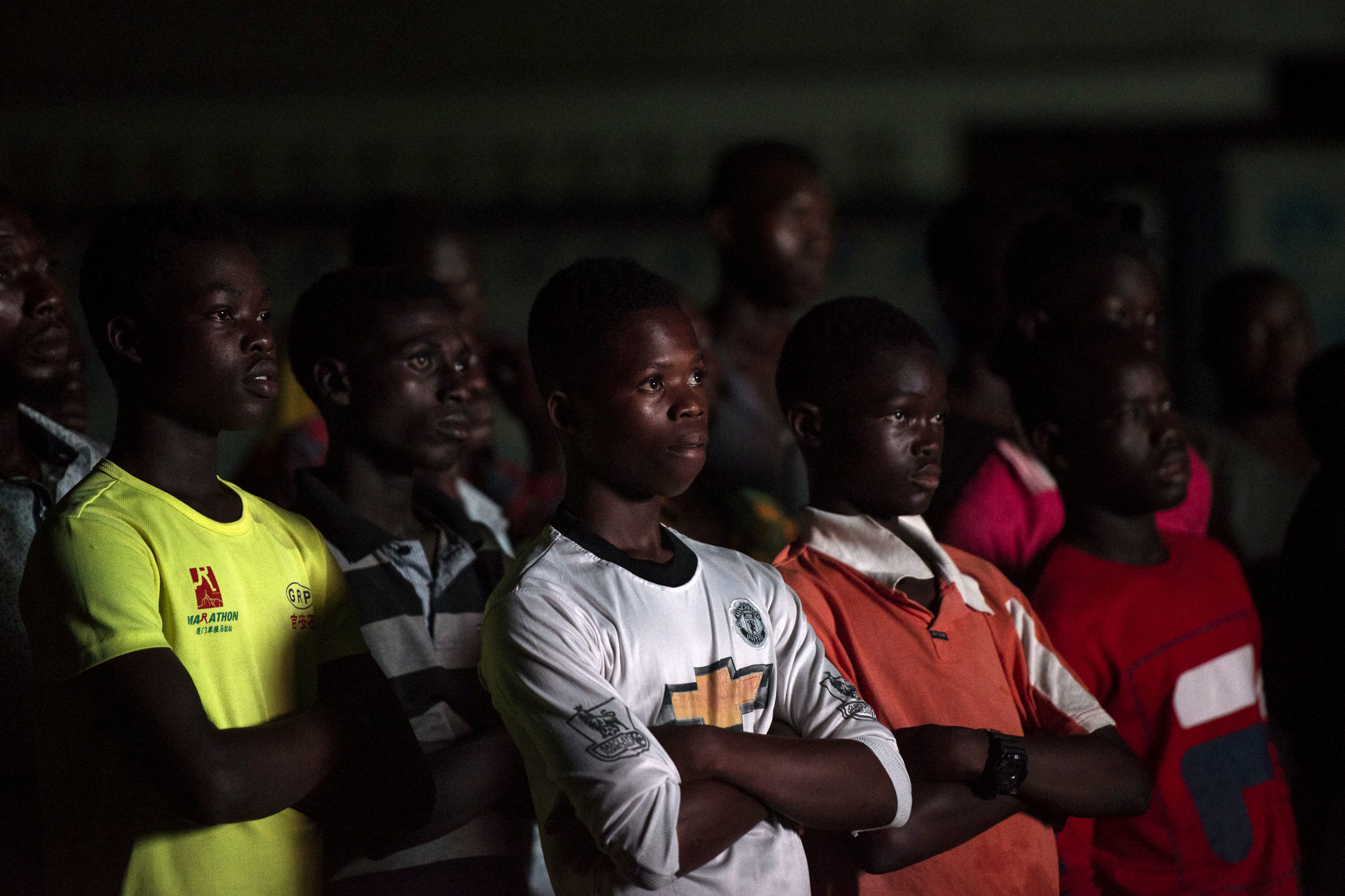 Against a dark background, a group of young boys with soccer jerseys on stand with their arms crossed as that watch a film out of the frame