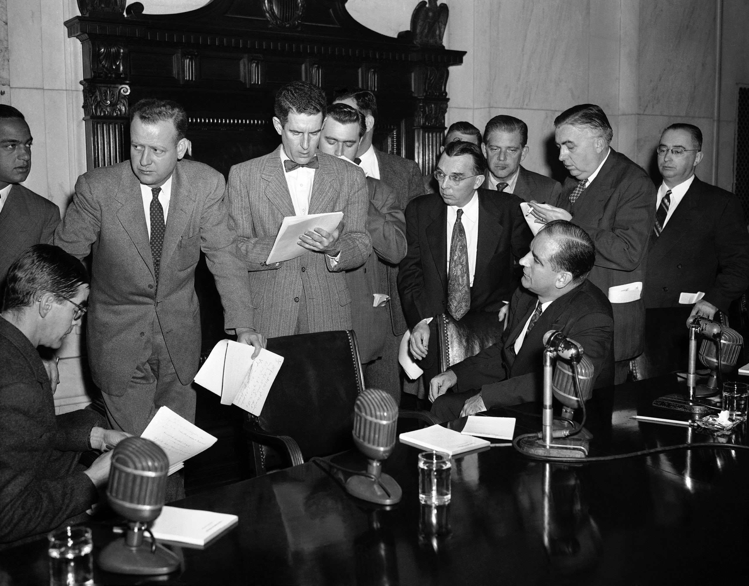 Joseph McCarthy sits at the right of a long table with four microphones on it. Surrounding McCarthy and the table, a heard of journalists with pens and notepads in hand stand over McCarthy