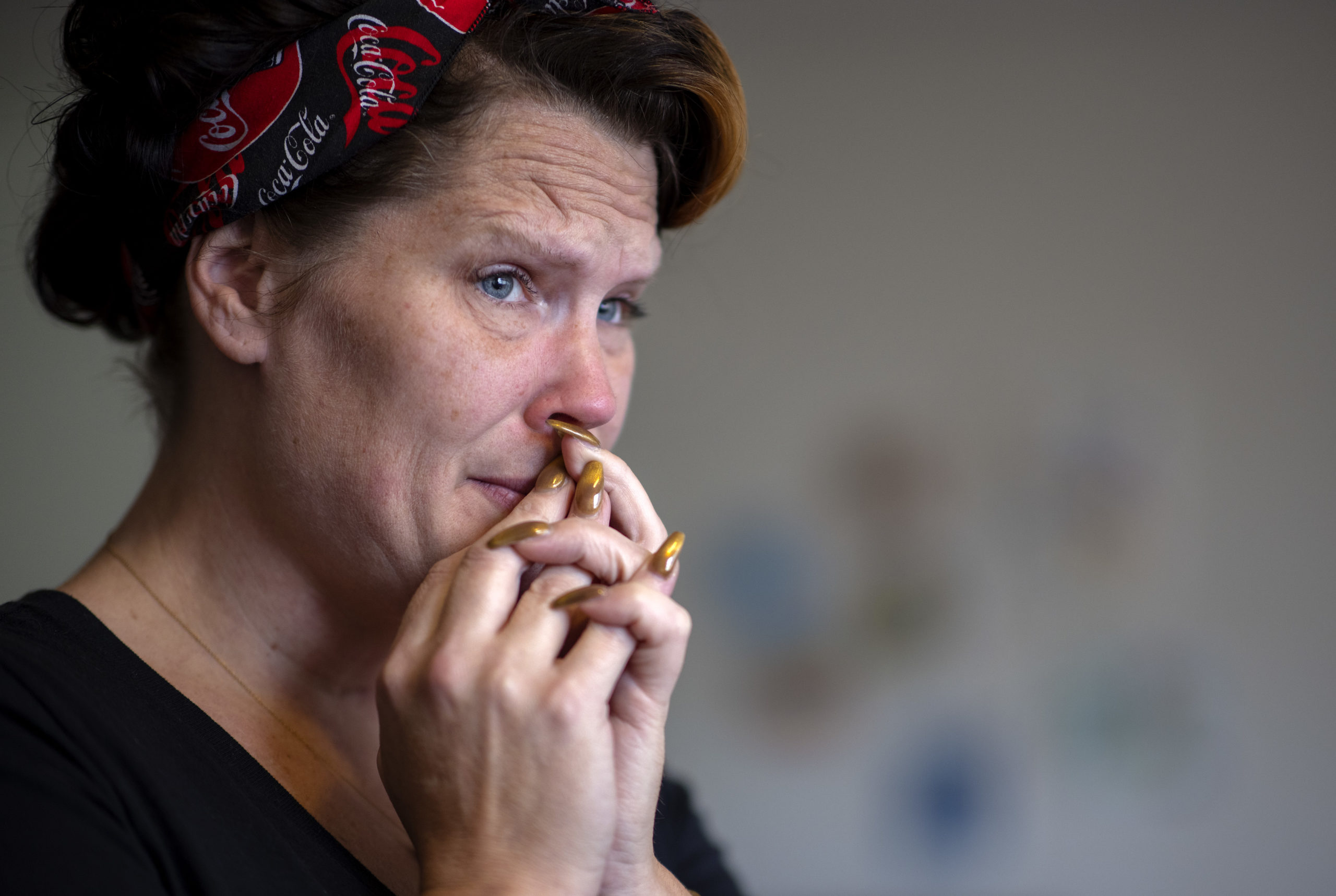 A side profile shot of Courteney Ross, where she stands left wearing a Coca Cola bandana headband and a black shirt. She folds her hands over her lips, wearing gold nail polish