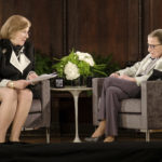 Against a brown curtain, Nina Totenberg, who wears a black dress and holds a stack of papers, interviews Ruth Bader Ginsberg on stage