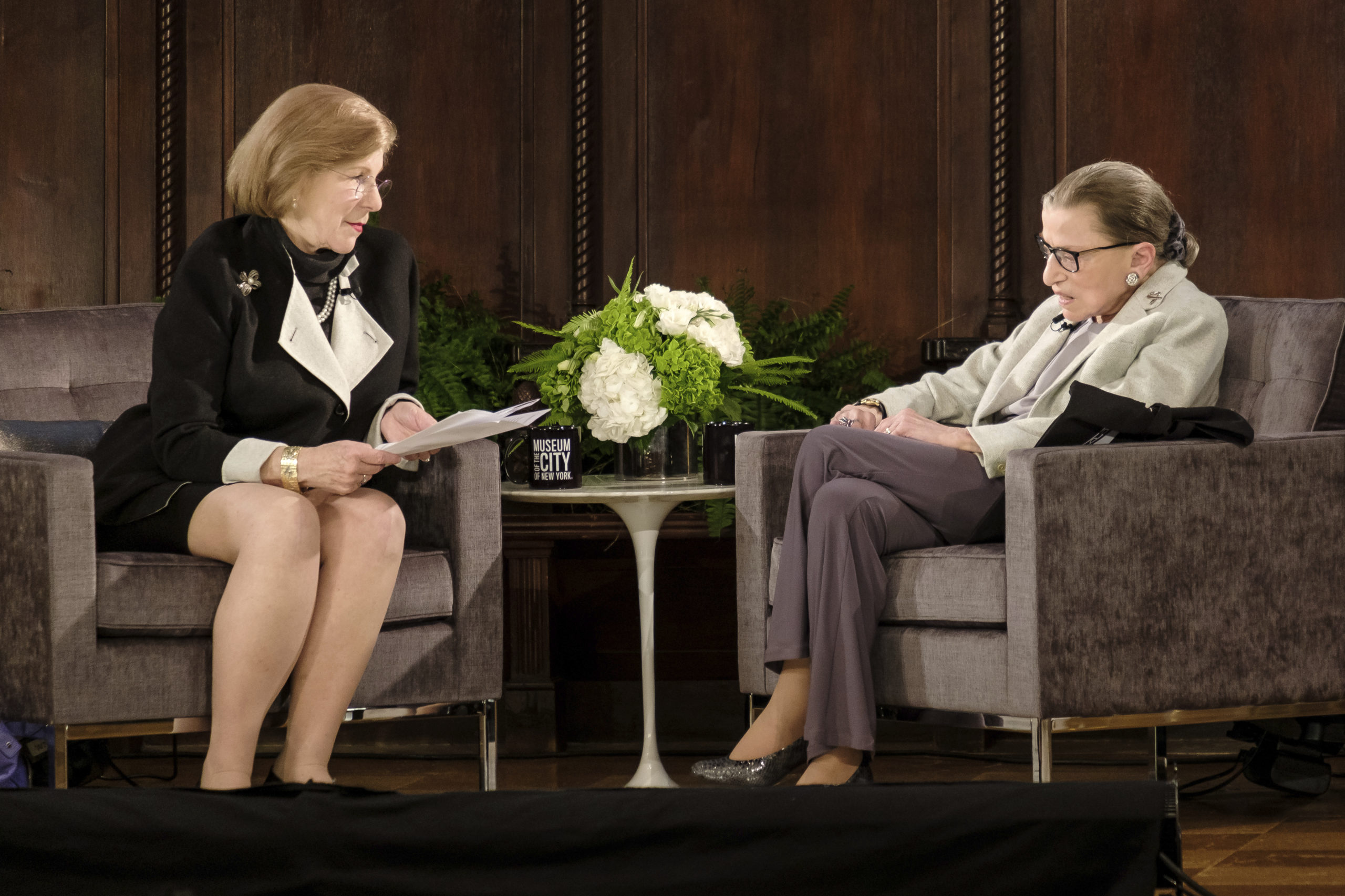 Against a brown curtain, Nina Totenberg, who wears a black dress and holds a stack of papers, interviews Ruth Bader Ginsberg on stage