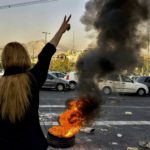 Standing to the left, a woman with her back facing the camera holds up a peace sign. In front of her, a fire blazes in the middle of the road