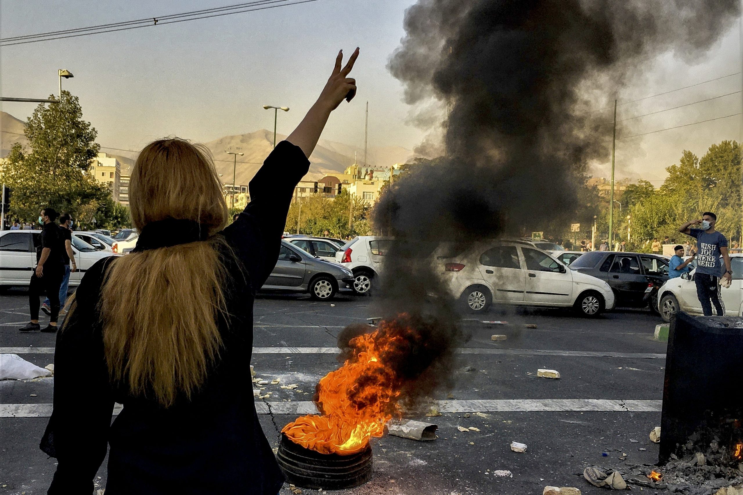Standing to the left, a woman with her back facing the camera holds up a peace sign. In front of her, a fire blazes in the middle of the road