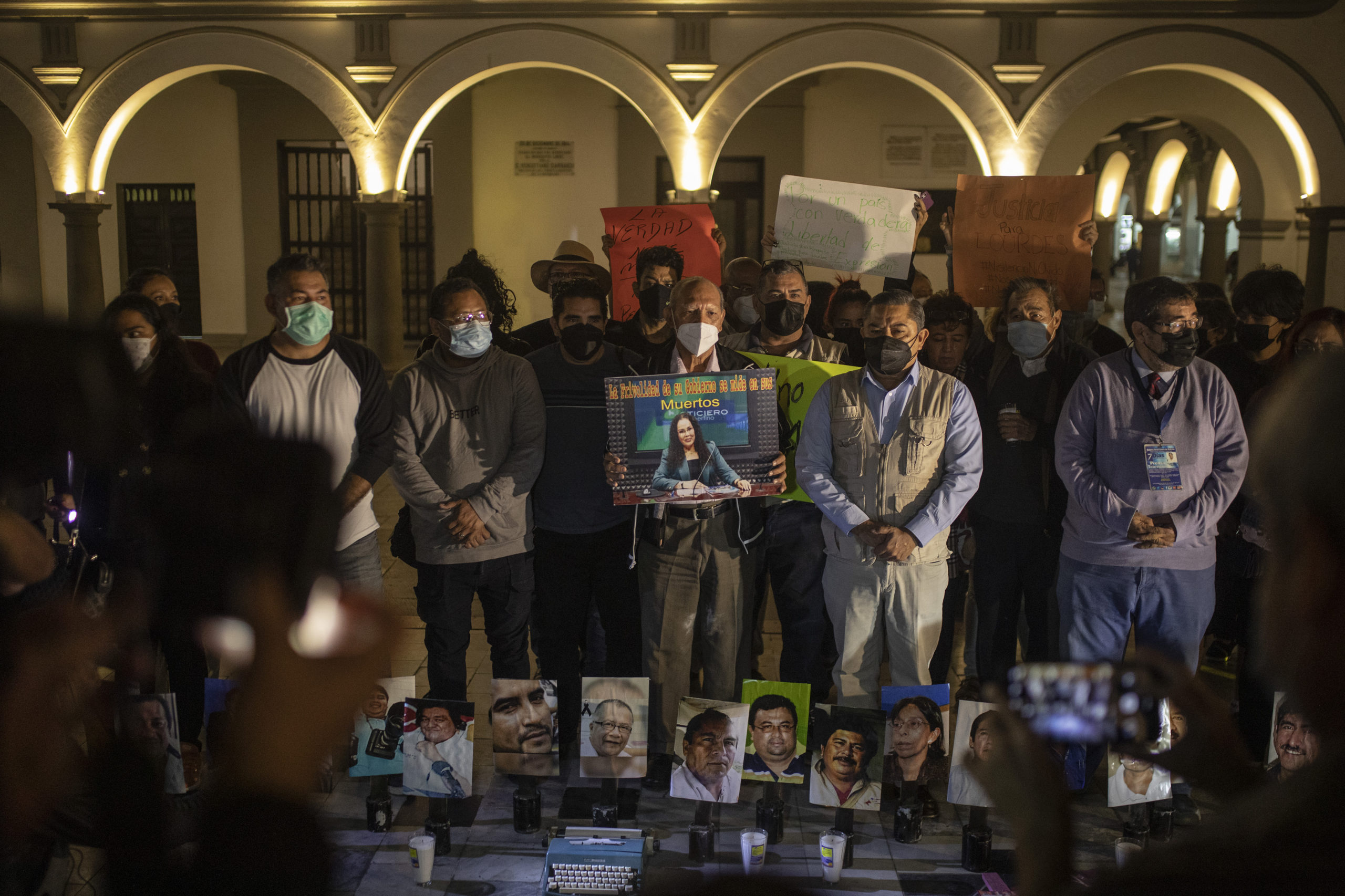 On the floor are over a dozen photos of journalists murdered, with candles surrounding them and a typewriter at the center. A line of people stand behind the photo vigil, some holding signs. The person in the center holds a sign that reads "La Frivolidad de su Gobierno se mide en sus Muertos"