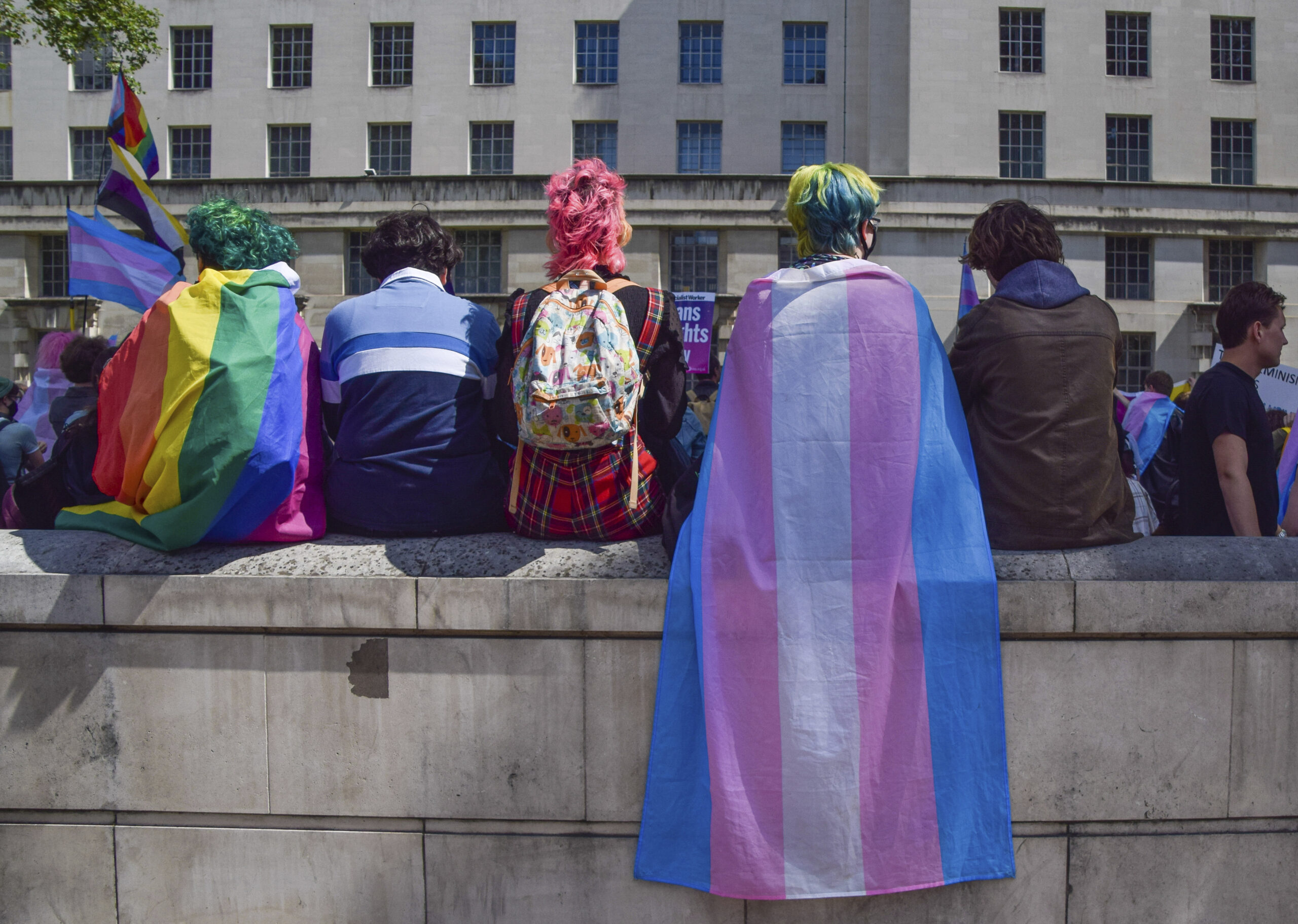 Protesters wrapped in pride and trans pride flags sit on a wall during the trans rights demonstration