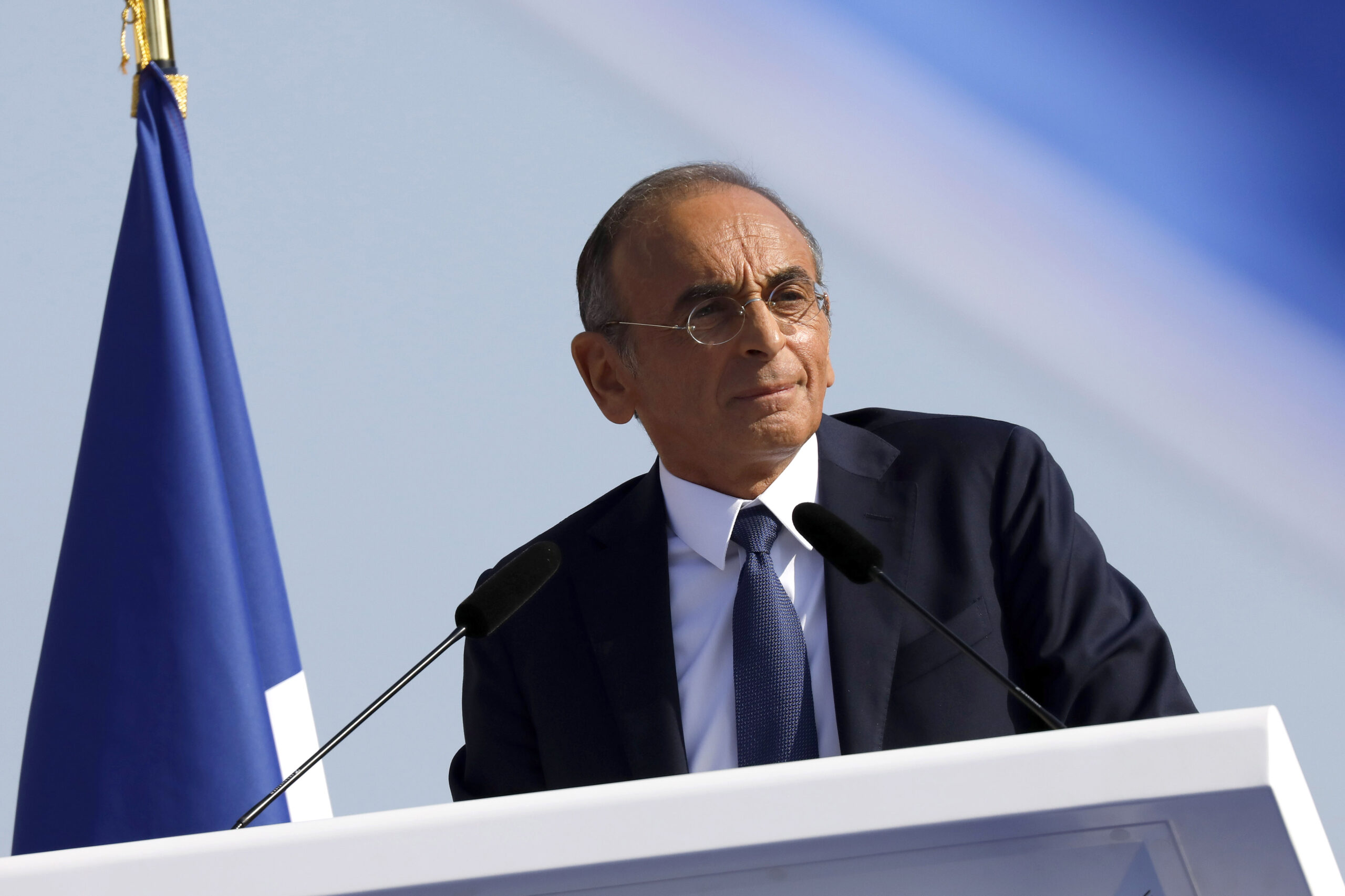 Zemmour, dressed in a black suit and blue tie, stands at a podium with a microphone