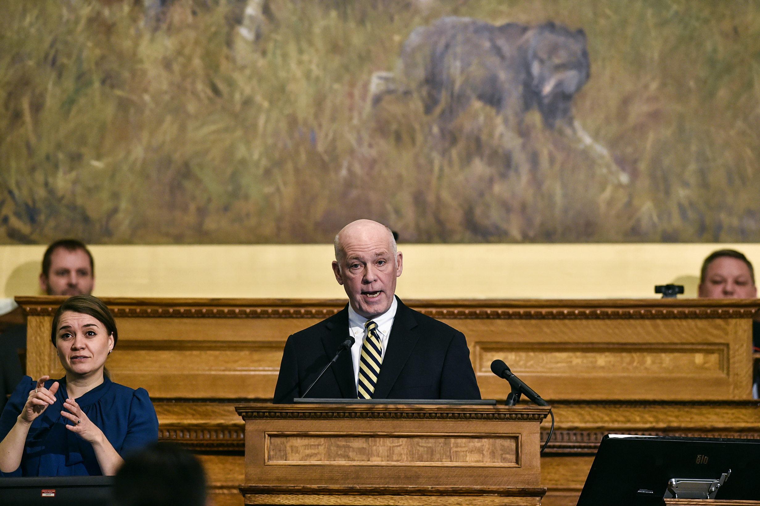 Gov. Greg Gianforte is standing at the podium of the Montana Senate/House of Reps building. He is wearing a black suit jacket, white shirt, and yellowish tie. An ASL interpreter is standing next to him.