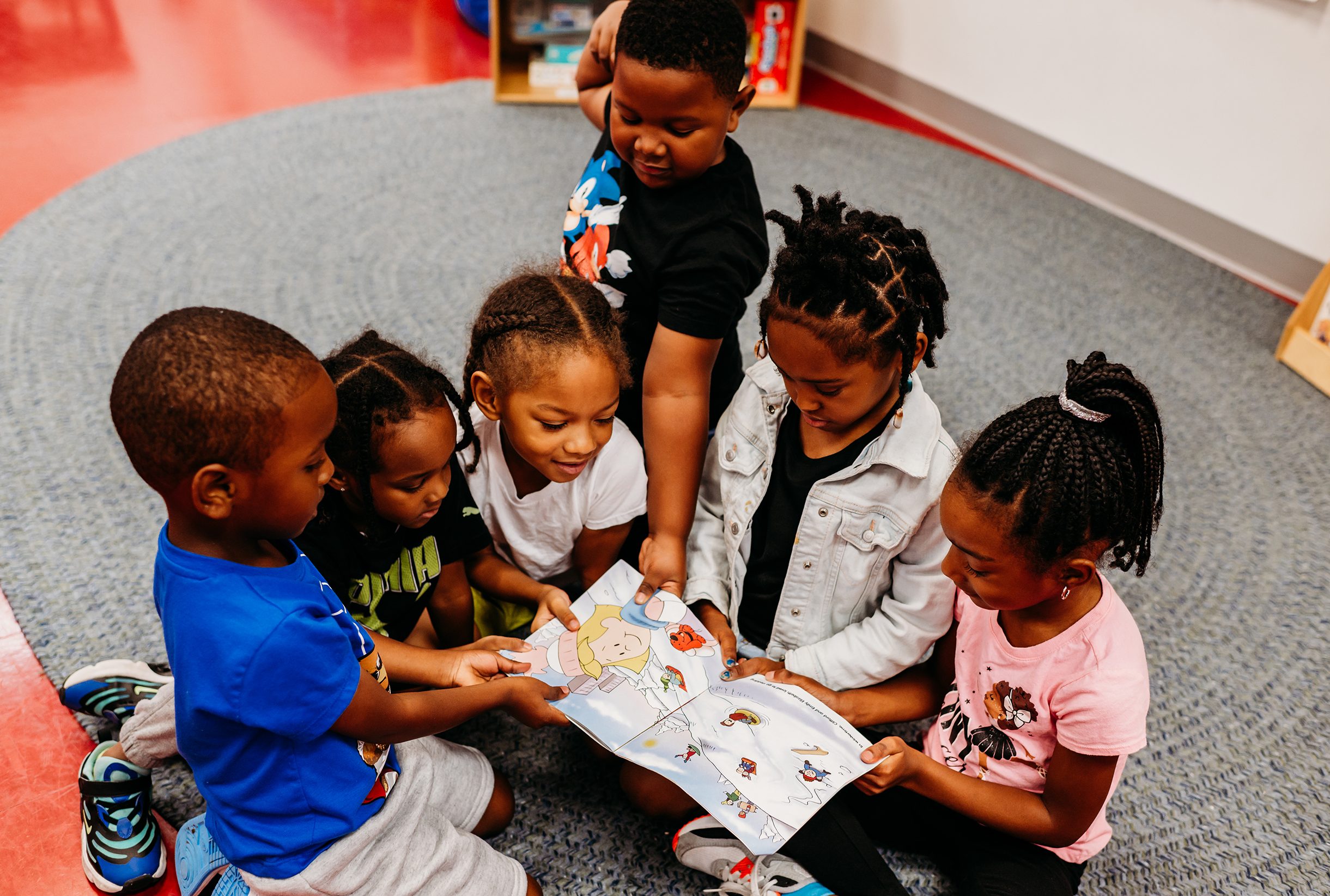 a group of toddlers/preschoolers are gathered around a picture book on the carpet of a daycare center.