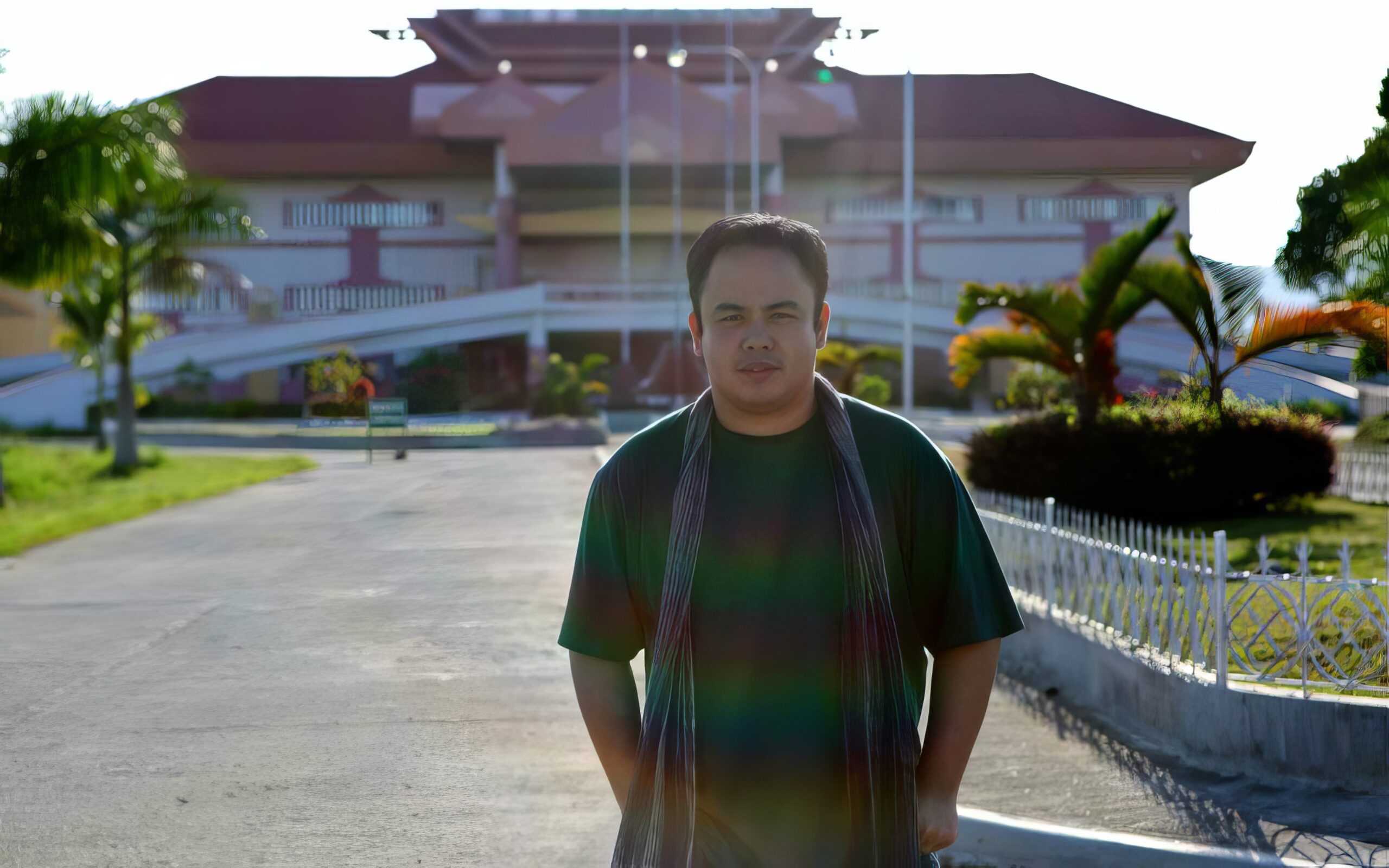 a man wearing a gray scarf, green t-shirt, stands in front of a capitol building in the Phillippines. The capitol building is a mixture of Spanish and indigenous styles of architectue, surrounded by palm trees.