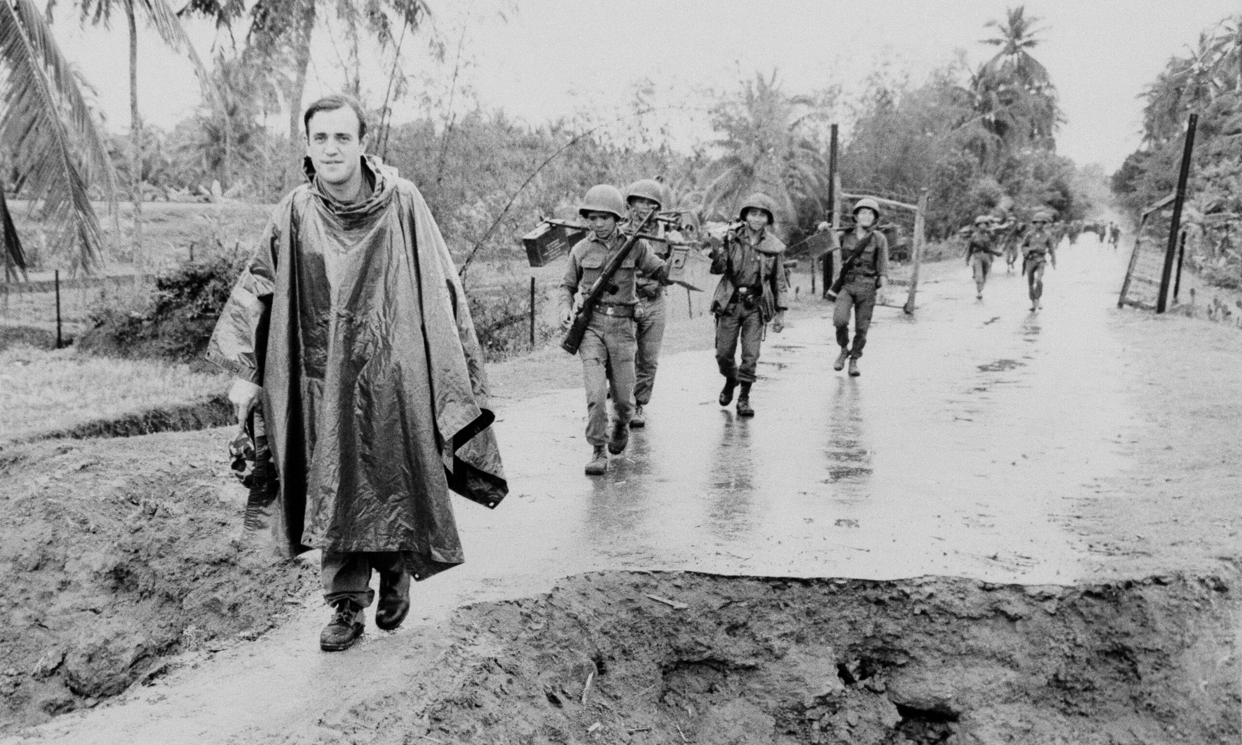 black and white photo of a man wearing an army poncho and trekking through the subtropical vietnamese jungle, there are soldiers trekking behind him